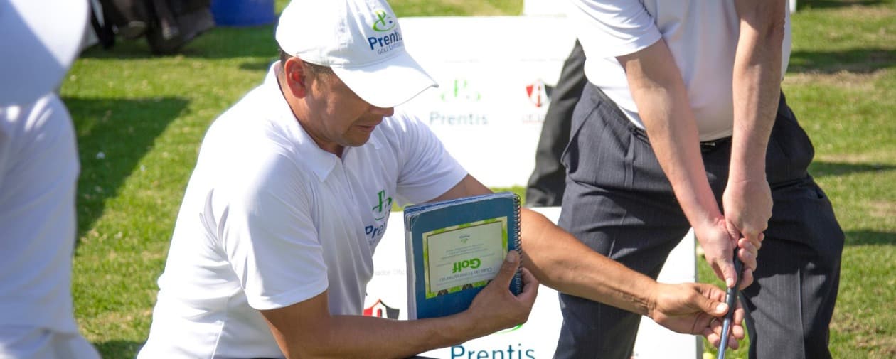 A Prentis Golf coach kneeling with the training guide in hand, adjusting a student’s grip on the club