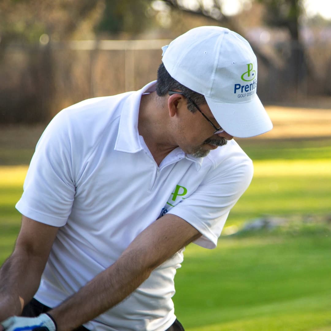 A Prentis Golf coach in a branded cap and polo turning through a swing on a sunny practice range