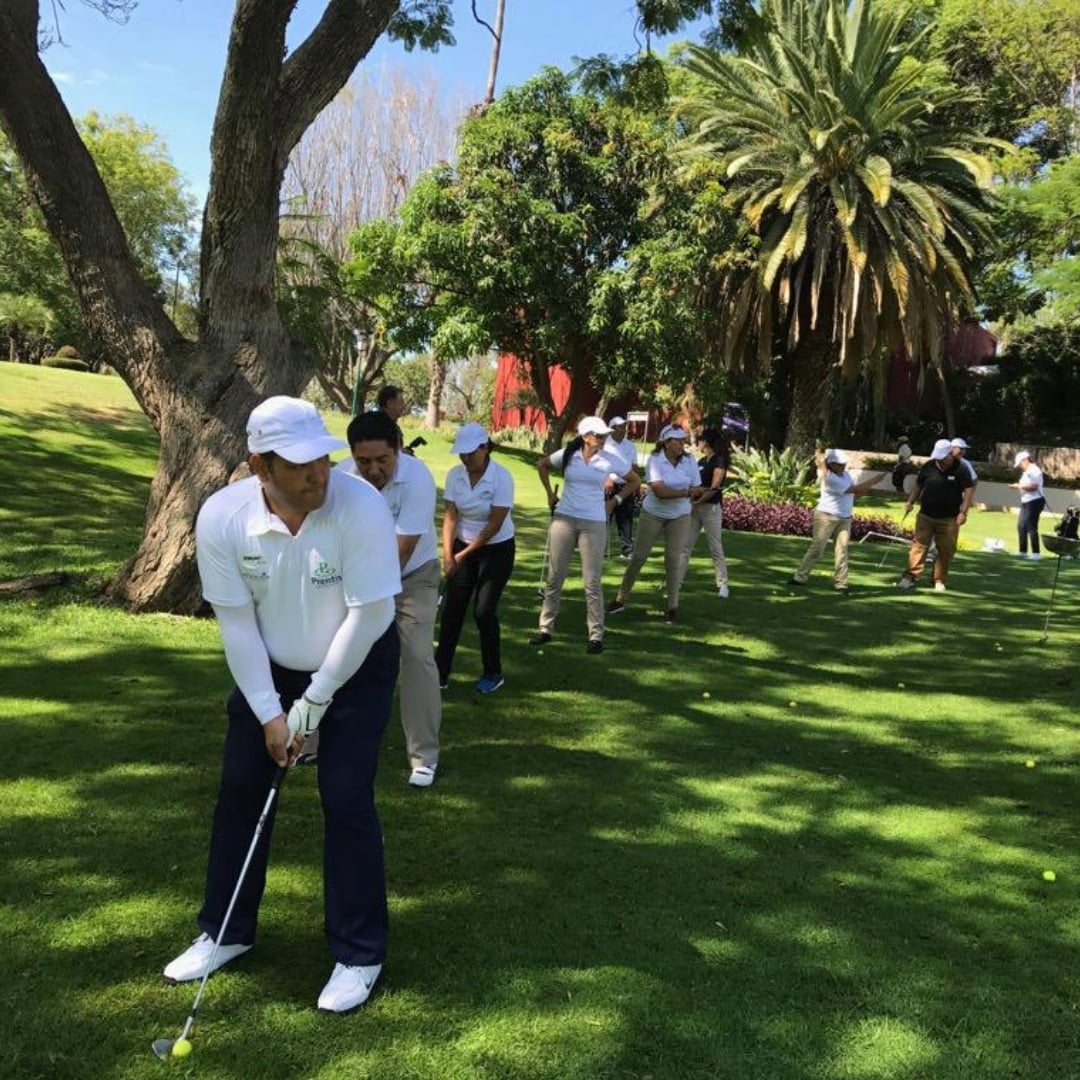 A line of Prentis Golf students in white caps and polos practicing chip shots on a tree-lined course